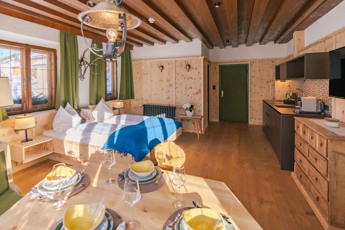 A wooden interior with a dining table set for two, a bed with a blue blanket, and a kitchen area with a TV.