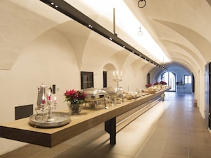 A long, narrow room with a high ceiling and arched walls, featuring a long table with various food service items and a decorative centerpiece. - Hotel Jagdschloss Resort (Silz)