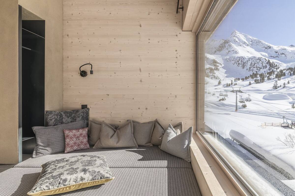 A room with a large window overlooking a snowy mountain landscape, a bed with patterned pillows, and a wooden wall.