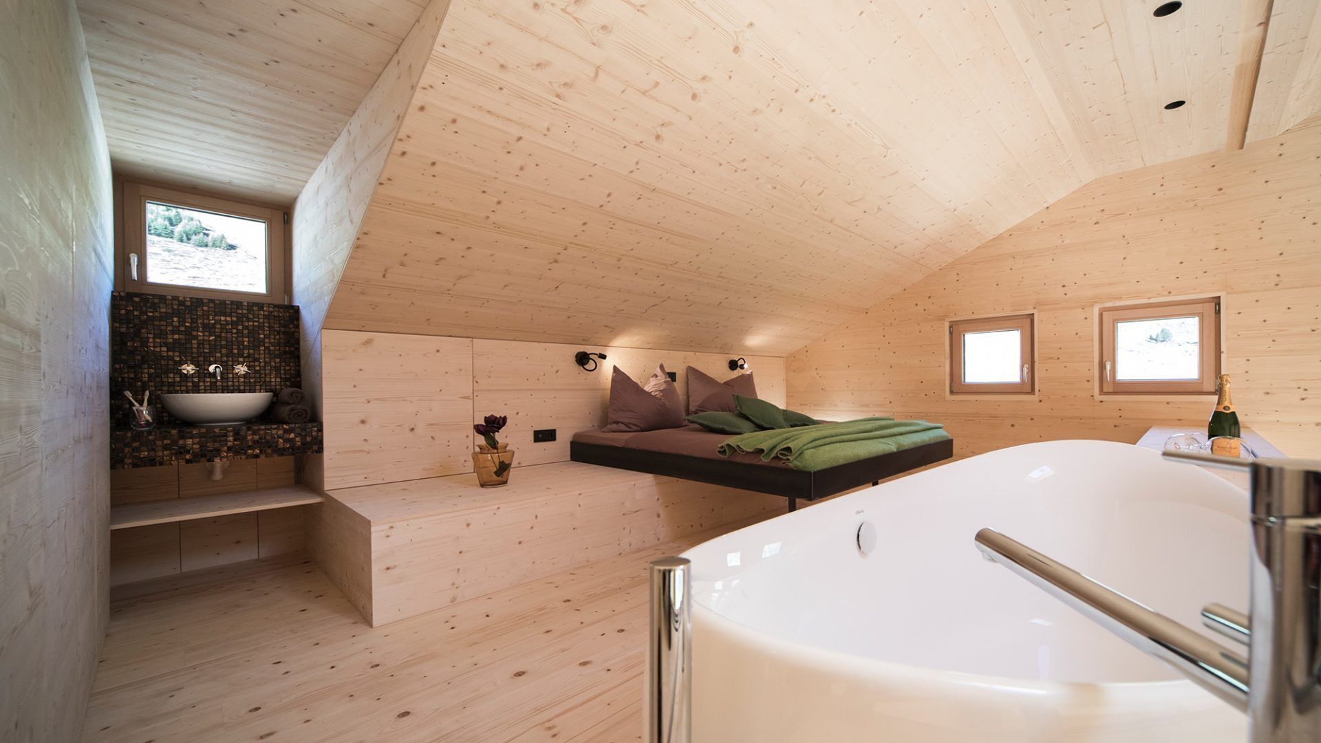 Un intérieur moderne en bois, doté d’une grande baignoire, d’une banquette intégrée avec des coussins, d’un petit coin lavabo et de deux fenêtres qui laissent entrer la lumière naturelle.