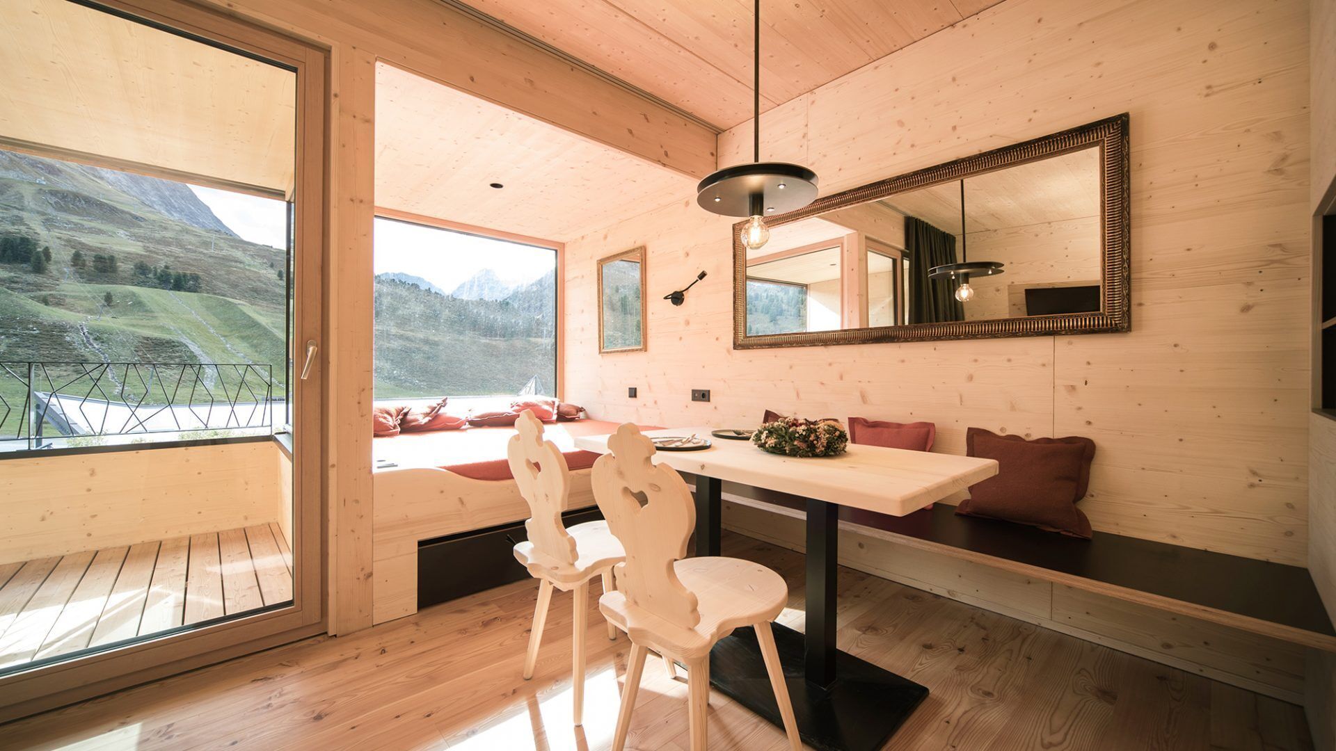 Un intérieur moderne avec des murs en bois, une grande fenêtre offrant une vue sur les montagnes, un coin repas avec une table et des chaises, et un banc agrémenté de coussins.
