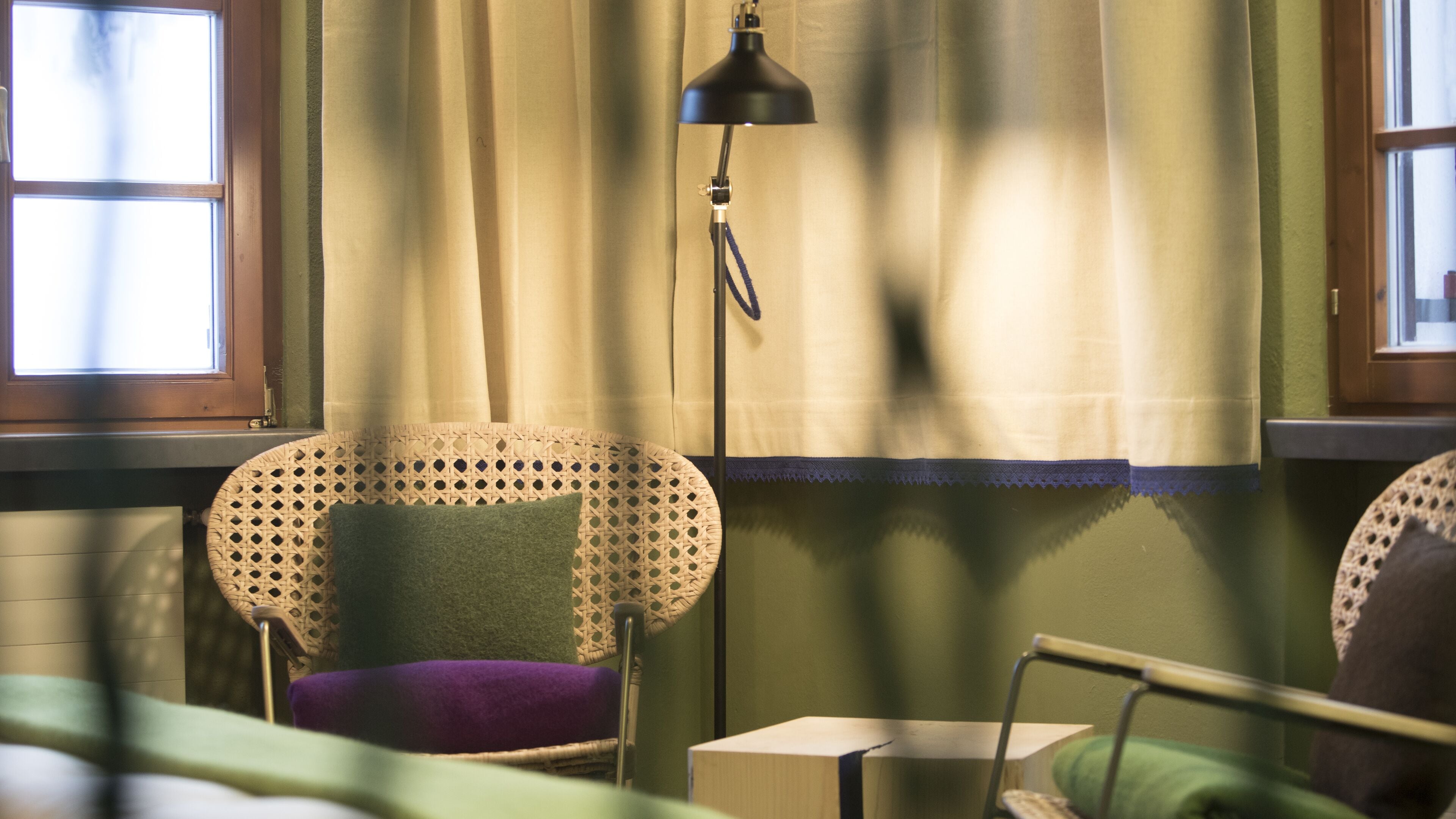 A room with a wicker chair, a small bedside table, a lamp, and a window with curtains.