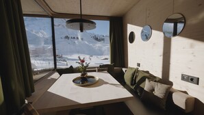 Een knus ingerichte kamer met een groot raam en uitzicht op een besneeuwde berg, een eettafel met een bloemenvaas, en een bank met kussens.