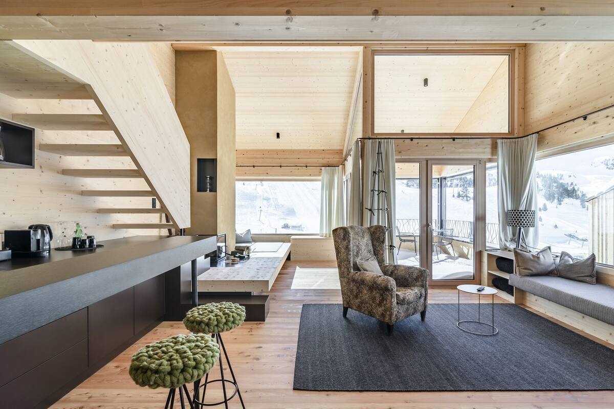 A modern interior with a wooden staircase, a bar area, a seating area with a sofa and armchair, and large windows offering a view of the outdoors.