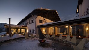 A snow-covered outdoor seating area with lounge chairs and a fire pit, adjacent to a building with large windows and a thatched roof. - Hotel Jagdschloss Resort (Silz)