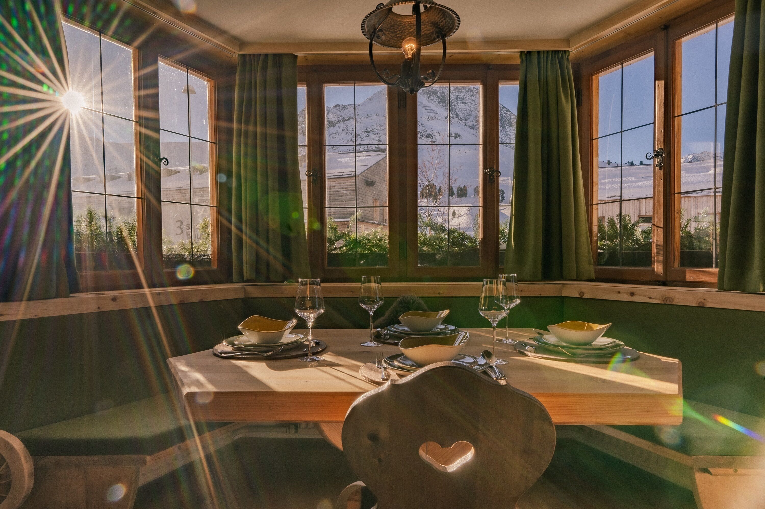 A dining area with a wooden table set for two, large windows with curtains, and a view of snow-covered mountains.