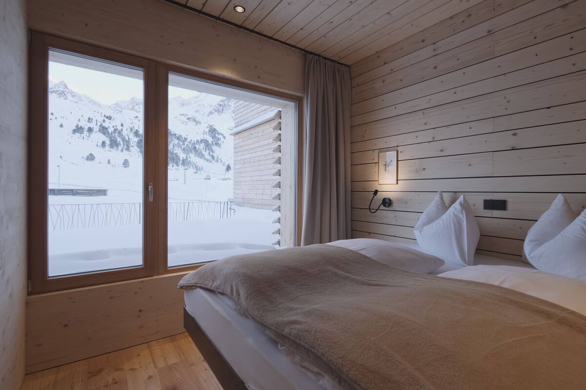 A bedroom with a bed, wooden walls, a window with a snowy mountain view, and a curtain.