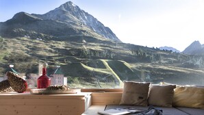 A room with a view of a mountain landscape, a wooden table with a bottle and pine cones, and a sofa with pillows. - Appartements Seenhaus (Silz)