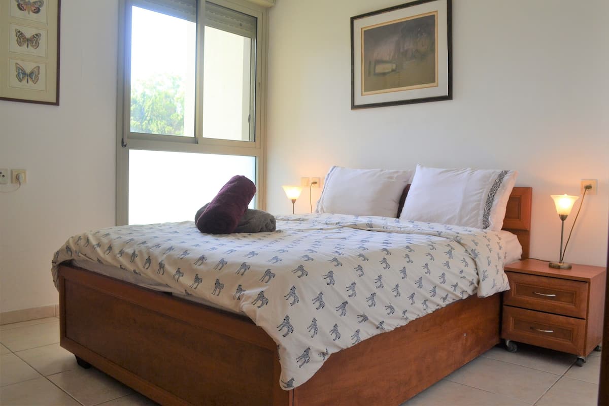 Une chambre à coucher avec un lit en bois, une fenêtre, une table de chevet avec une lampe et des œuvres d’art encadrées accrochées aux murs.