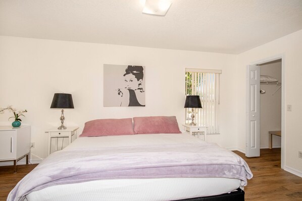 Une chambre à coucher comprenant un lit, deux tables de chevet, une commode, une fenêtre avec des stores, une porte et une œuvre d’art accrochée au mur.
