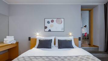 Habitación de hotel con cama, mesitas de noche, una obra de arte en la pared y una puerta que da a otra habitación.