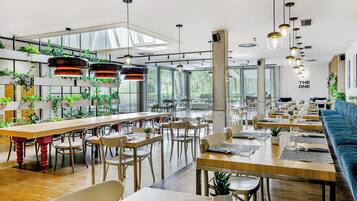 Un comedor amplio con mesas largas, sillas, lámparas colgantes y plantas en macetas.