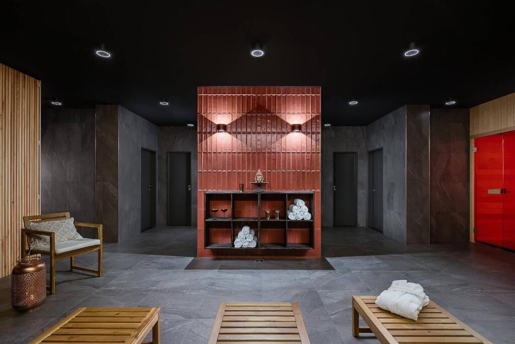 A modern lobby with a central wooden bench, a seating area with a chair, a red wall with shelves, and a red door.