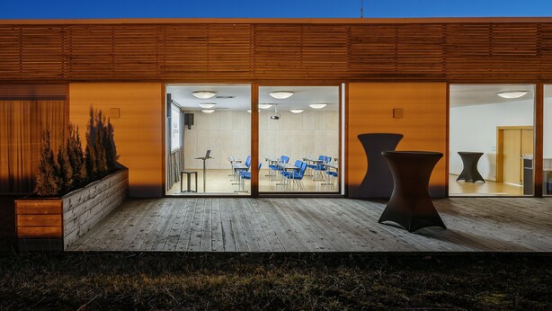 Un edificio moderno con fachada de madera, entrada de vidrio y un área de estar con mesas y sillas.
