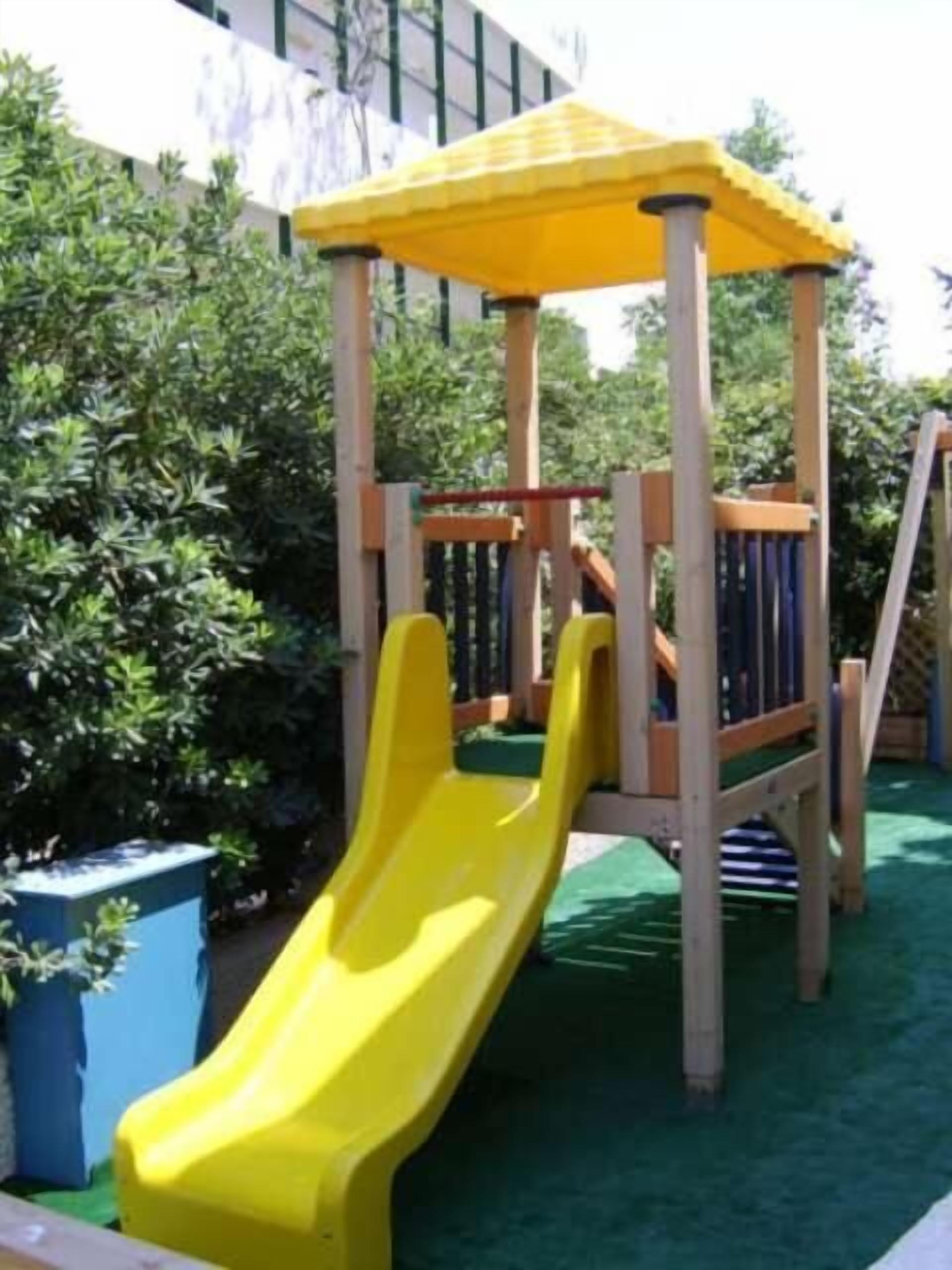 A wooden play structure with a yellow slide and a climbing area, surrounded by greenery.