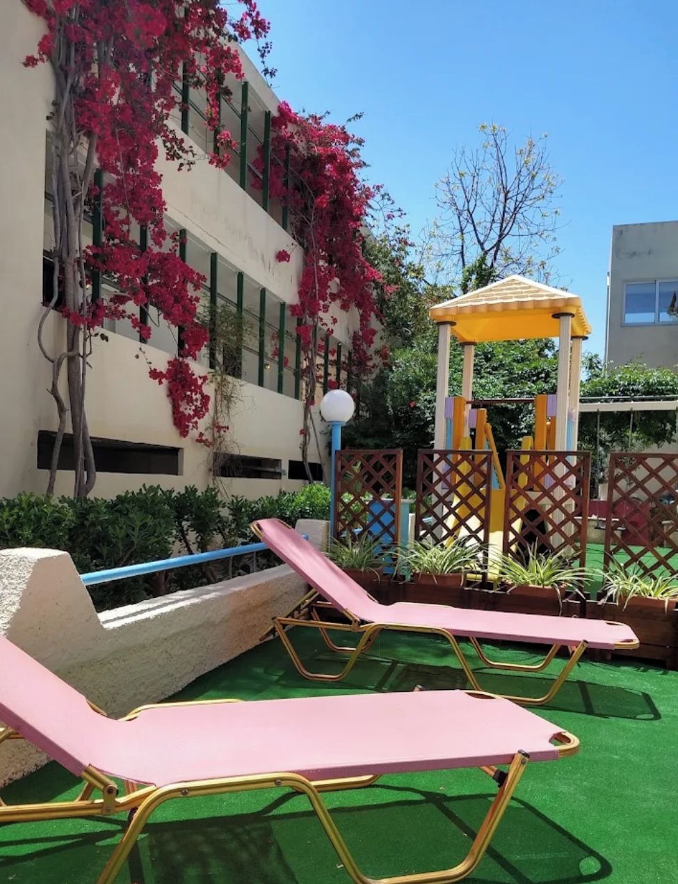 Une cour avec des chaises longues, une aire de jeux pour enfants et un bâtiment recouvert de plantes grimpantes.