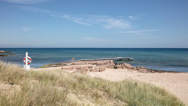 Une plage avec un panneau indiquant la présence d’une bouée de sauvetage, un rivage rocheux et une jetée qui s’avance dans l’eau.