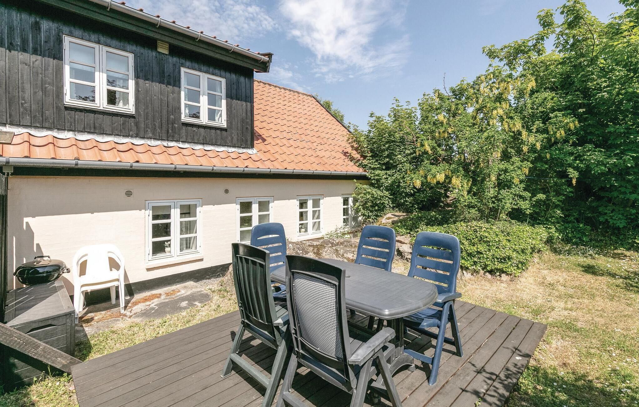 A wooden deck with a table and chairs, a house with a red-tiled roof, and a garden with trees and shrubs.