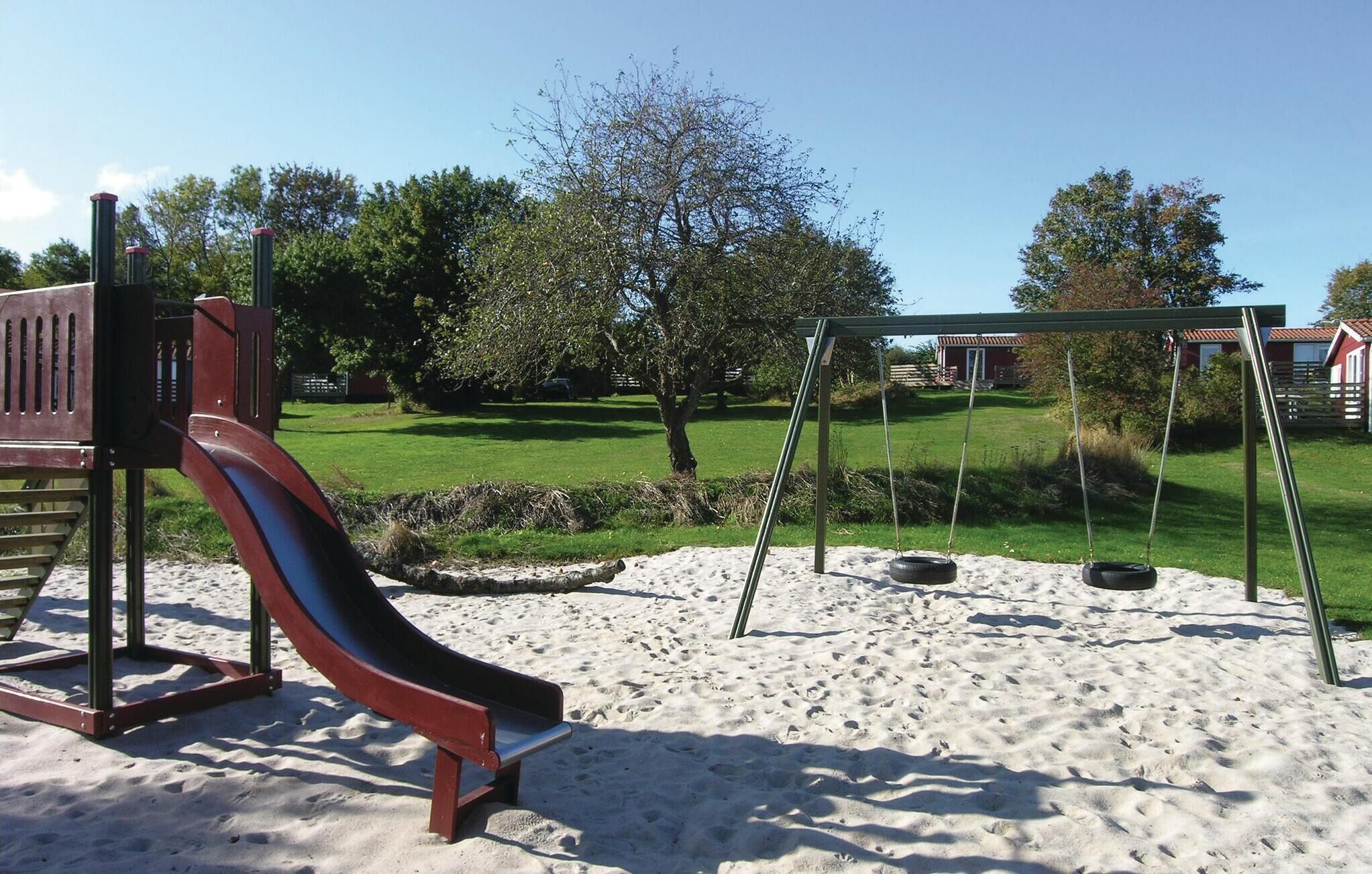 A playground with a slide and swings on a sandy surface, surrounded by grass and trees.