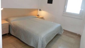 Un lit simple avec une couvre-lit blanche, une lampe de chevet, une petite table de nuit et une fenêtre avec des stores.