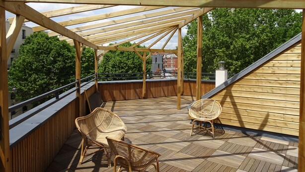 Una terraza de madera con un pérgola, dos sillas de mimbre y vistas a árboles y edificios.