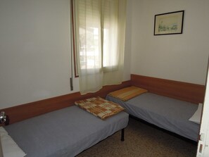 Quarto com duas camas de solteiro, janela com cortinas translúcidas, quadro na parede e porta de madeira.