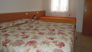 Une chambre à coucher avec un lit orné d’un motif corail et de coquillages, une lampe de chevet et une fenêtre avec des rideaux légers.
