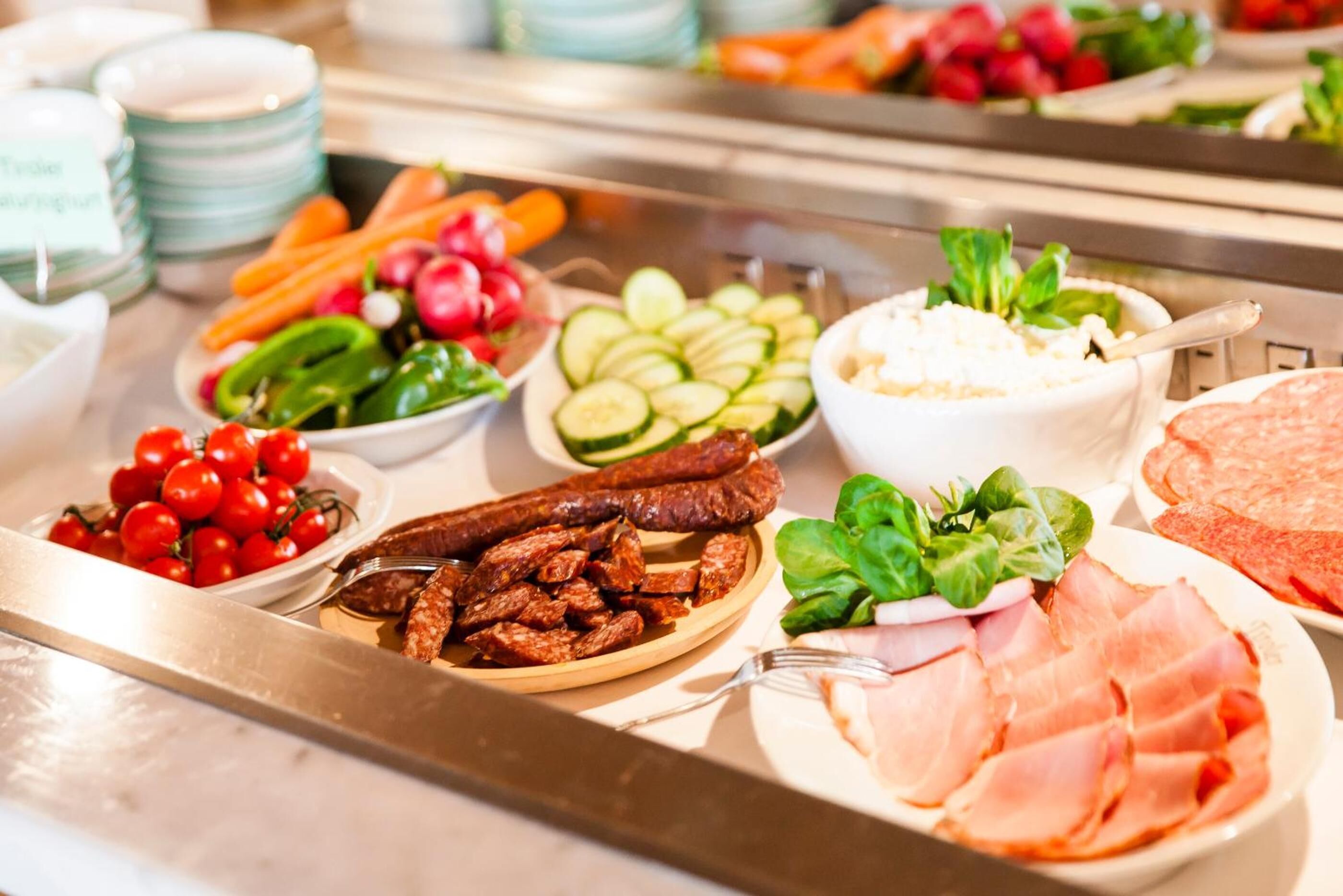 A buffet with various food items including sliced cucumbers, cherry tomatoes, sliced sausages, and leafy greens.