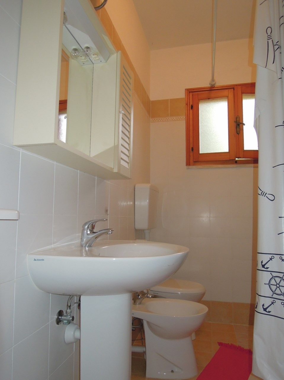 A bathroom with a white sink, a toilet, and a bidet. There is a mirror above the sink and a wooden-framed window above the toilet.
