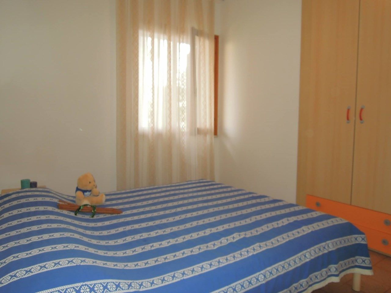 Un dormitorio con cama, un osito de peluche, una ventana con cortinas y un armario.