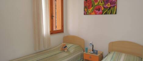 Un dormitorio con dos camas individuales, una ventana con cortinas, una mesita de noche con lámpara y un cuadro en la pared.