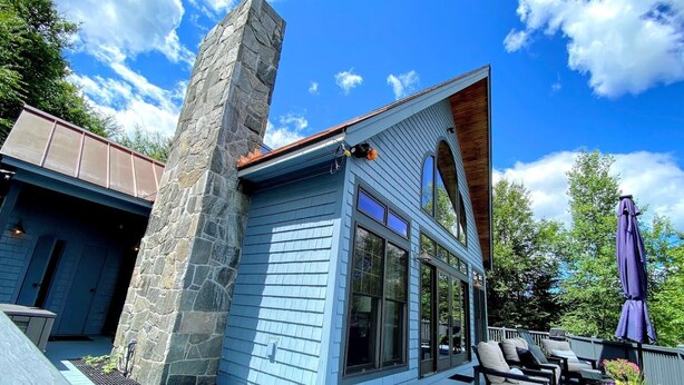 Une maison moderne avec une cheminée en pierre, de grandes fenêtres et une terrasse aménagée avec du mobilier d’extérieur.
