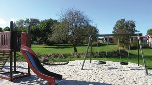 Een speeltuin met een glijbaan en schommels op een zandbodem, omringd door gras en bomen.