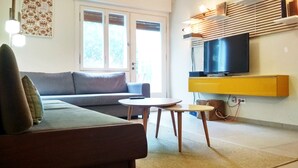 Uma sala de estar com um sofá, uma mesa redonda pequena, uma televisão sobre uma estante de madeira e uma janela com persianas.