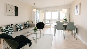 A modern living room with a sofa, coffee table, and chairs. There is a dining area with a table and chairs. The room has large windows and a view of the outside. - Lovely apartment in Allinge (Allinge)