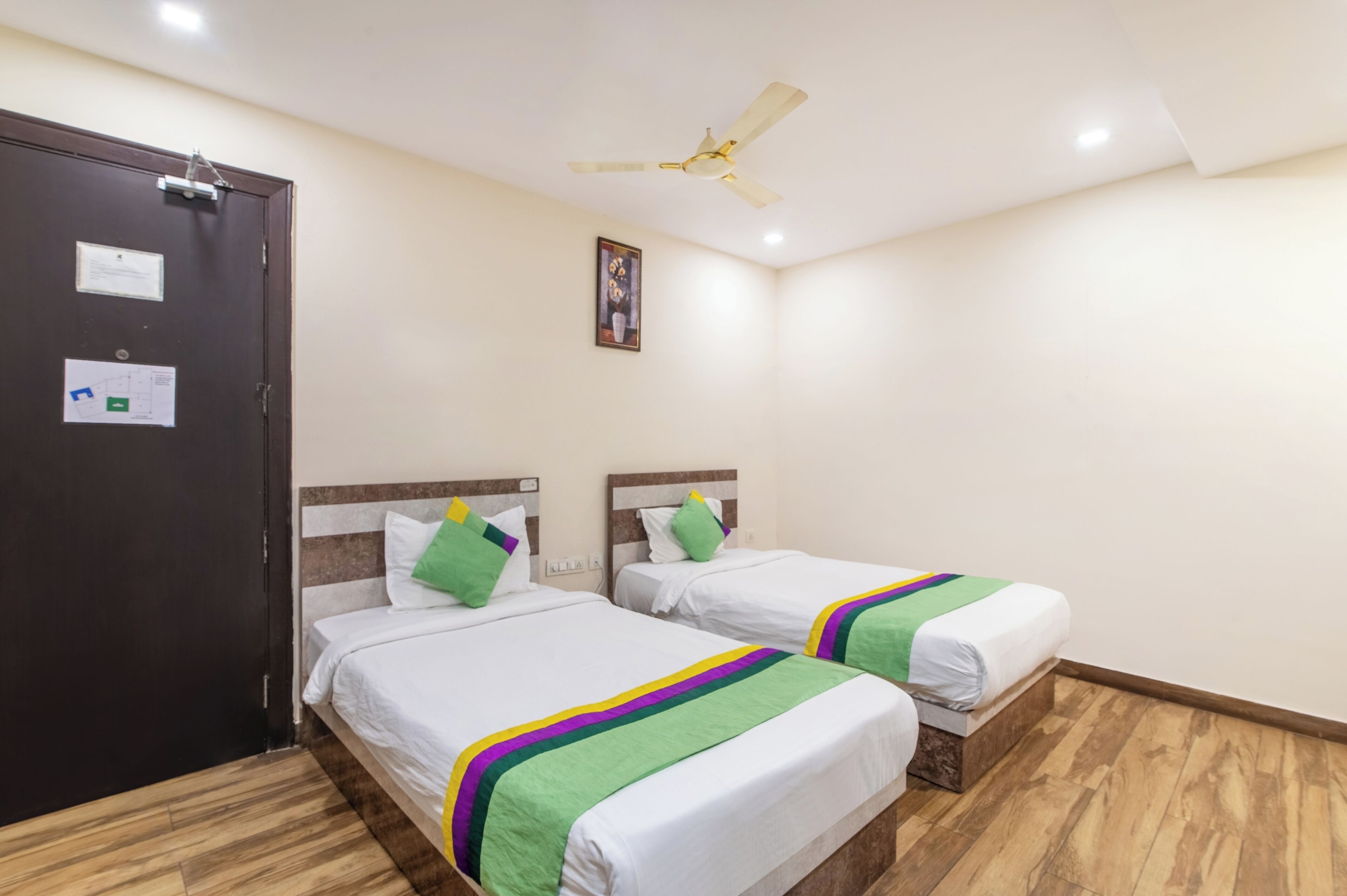 Une chambre d’hôtel avec deux lits, un ventilateur de plafond, une porte avec une serrure et un tableau encadré au mur.