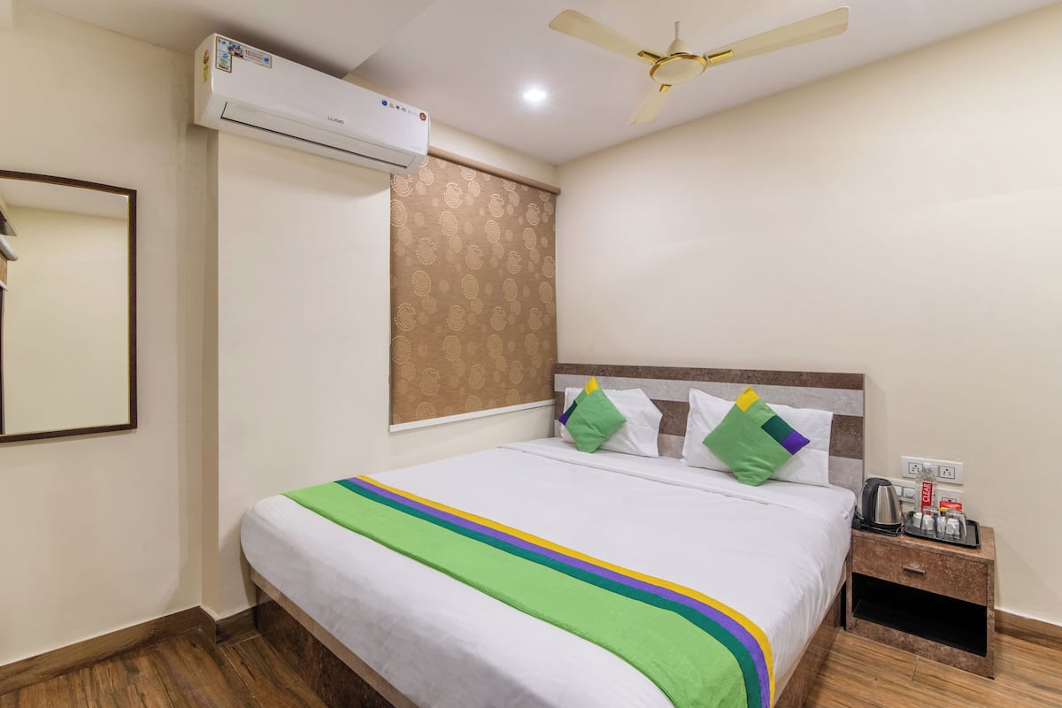 A hotel room with a bed, an air conditioning unit, a ceiling fan, a nightstand with a kettle, and a power outlet.