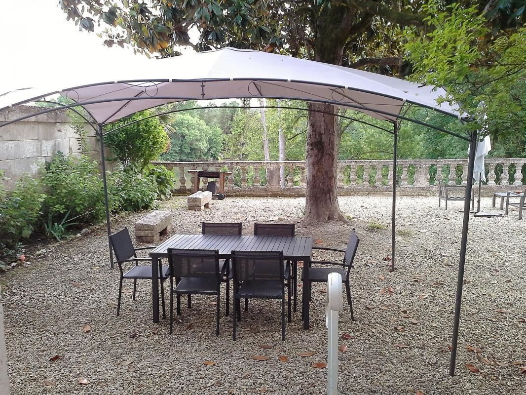 An outdoor patio with a table and chairs under a canopy, surrounded by gravel and greenery.