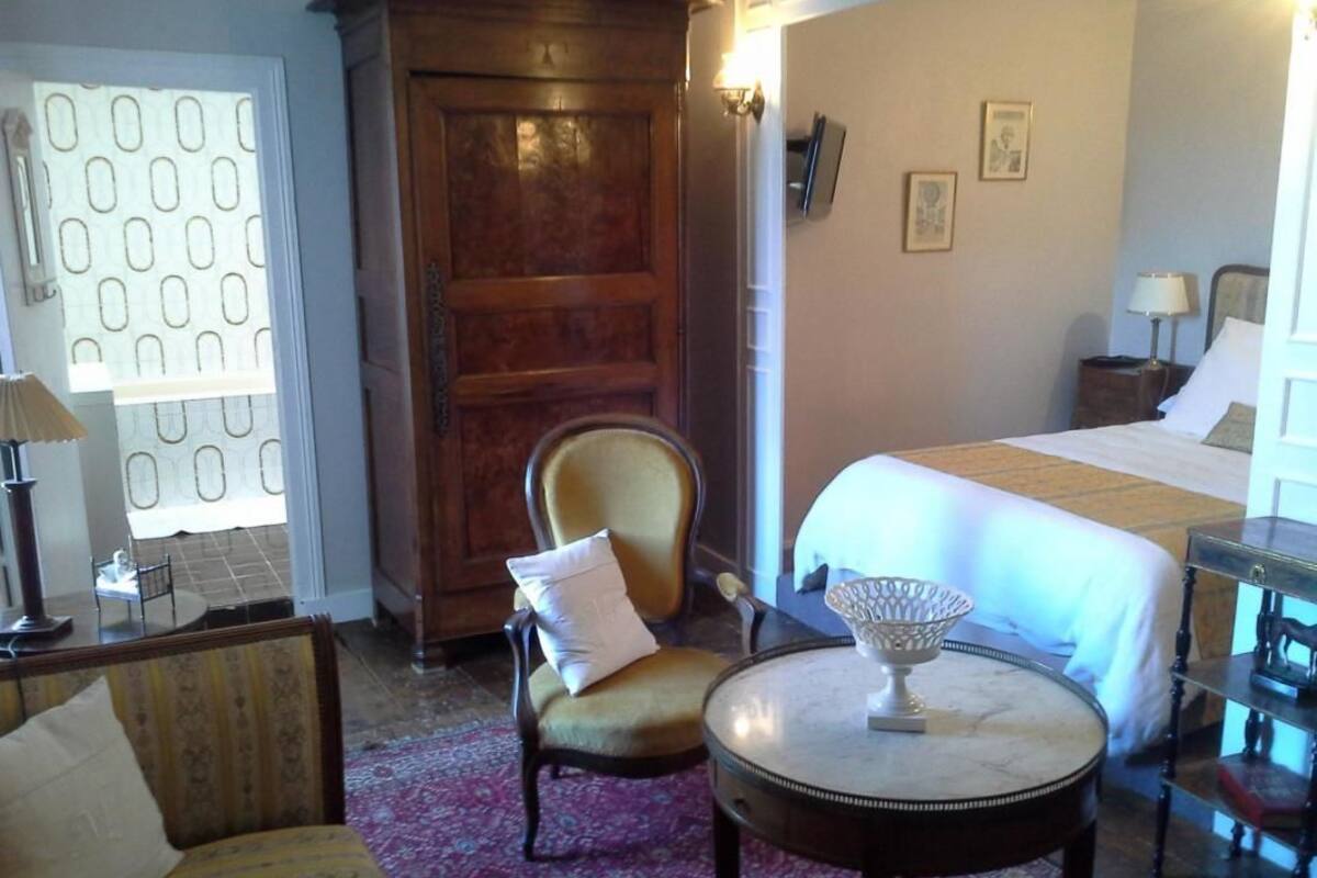 A hotel room with a bed, a chair, a small table, a nightstand, and a door with a patterned curtain.