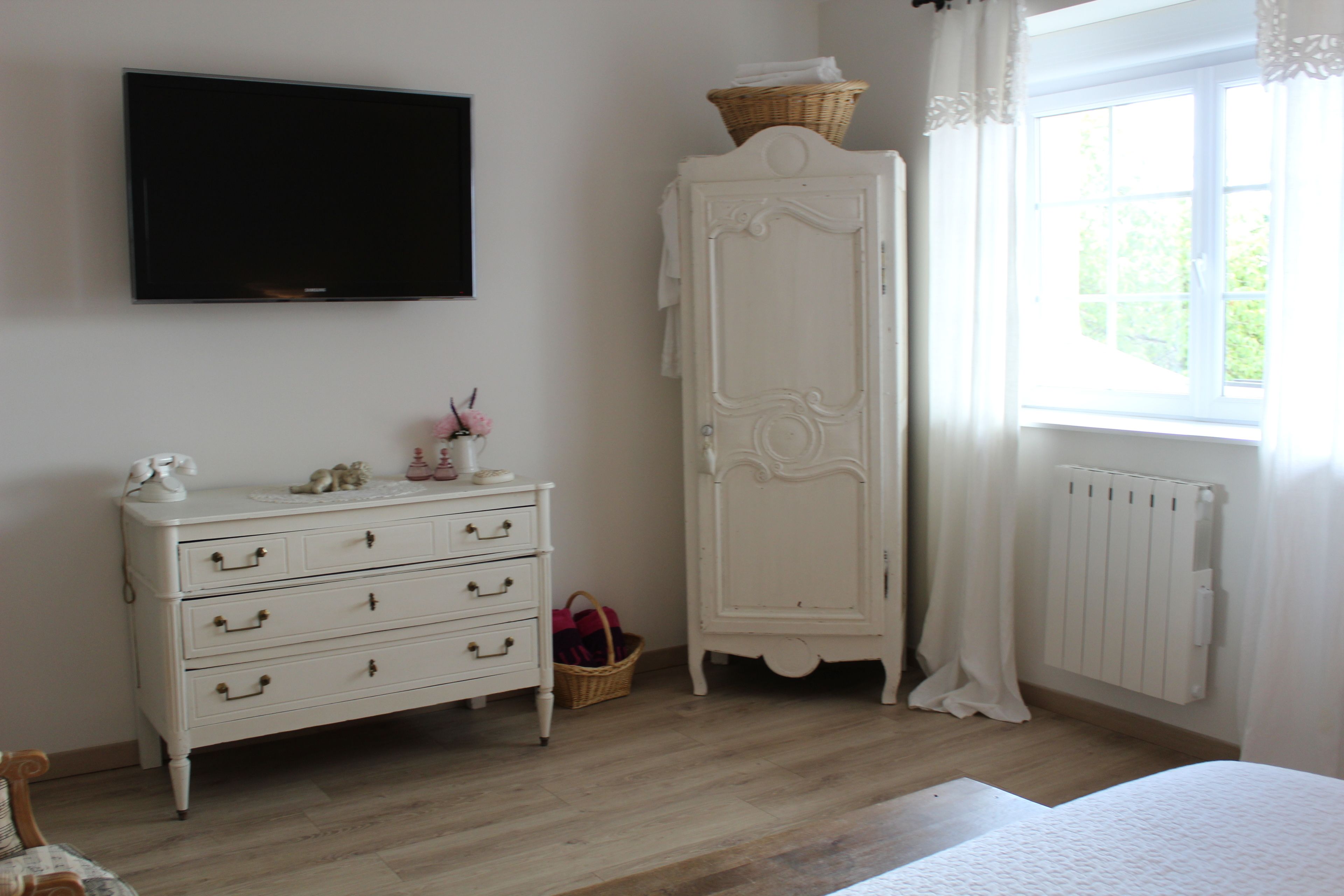 A bedroom with a white dresser, a television mounted on the wall, a window with curtains, and a radiator.