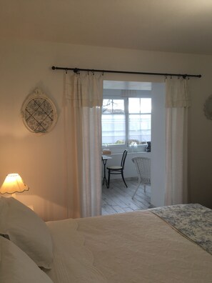 Quarto com cama, mesa e cadeira, em ambiente claro com vista para o exterior.