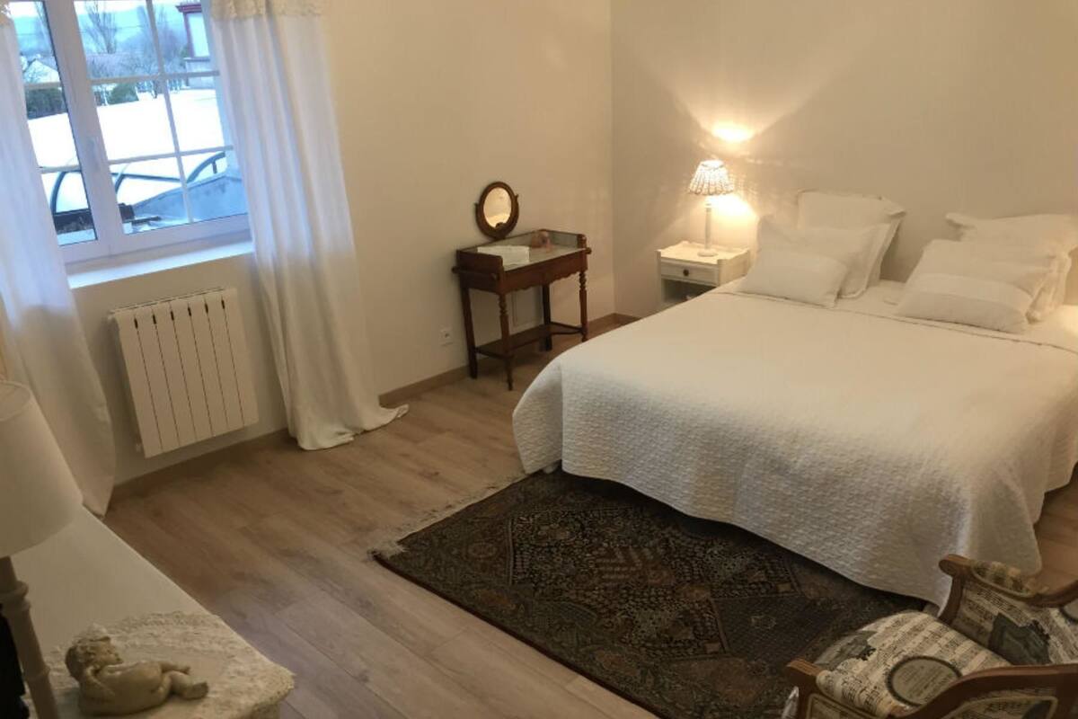 Une chambre à coucher comprenant un lit, une table de chevet, une lampe, une fenêtre avec des rideaux, un radiateur, un tapis et une chaise.