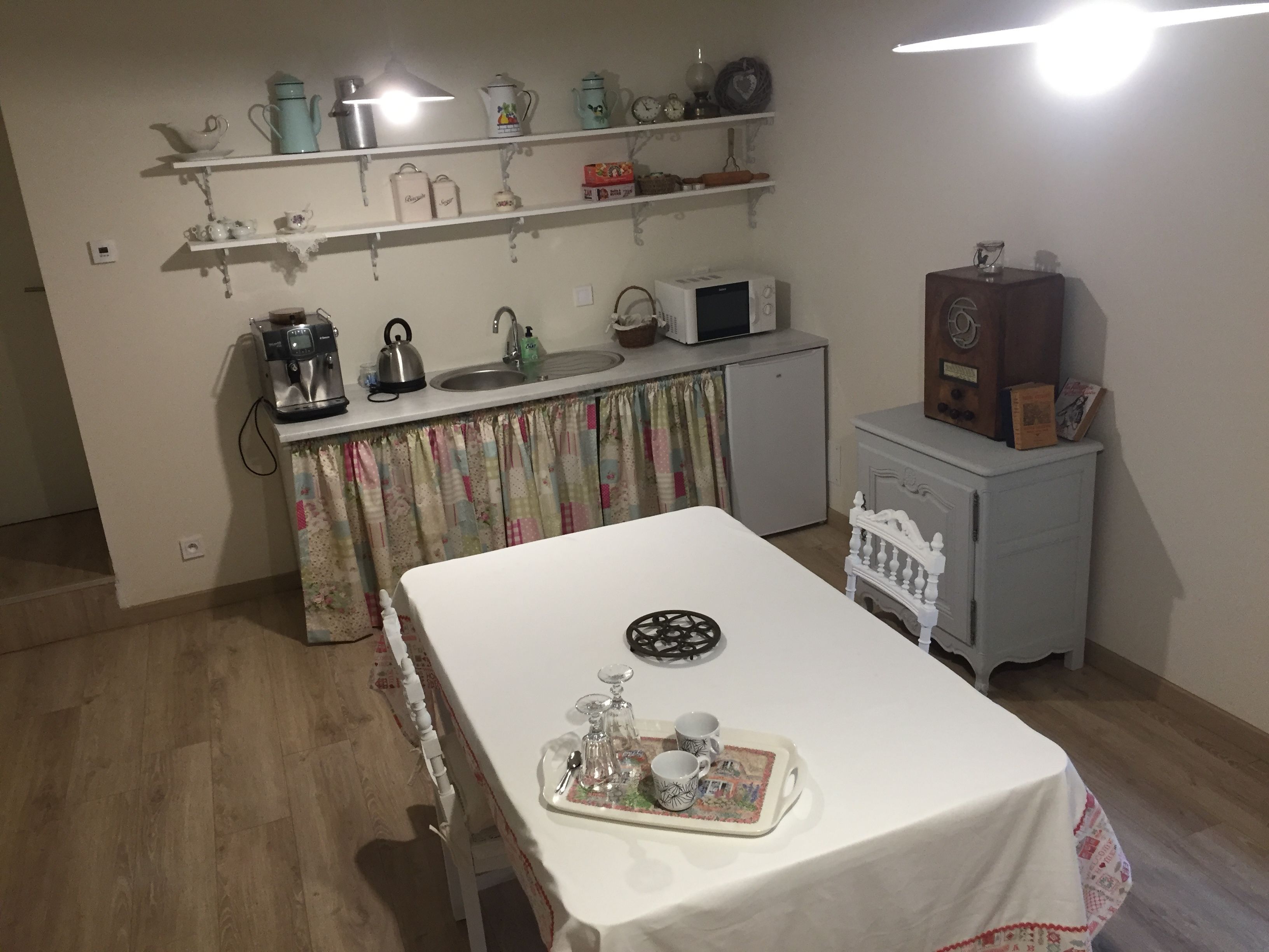 A compact kitchen with a microwave, kettle, and coffee maker on the countertop. A small refrigerator is visible, and a table with a tray of cups and a teapot is set up in the room.