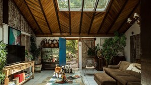 A spacious living room with a high ceiling, wooden beams, a large TV, a sofa, and various plants. - Pousada Casa do Ceo (Imbituba)