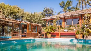 A pool area with a wooden building, a covered seating area, and a large window overlooking the pool. - Pousada Casa do Ceo (Imbituba)