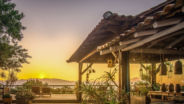 A wooden deck with a pergola, potted plants, and a view of the sunset over mountains. - Pousada Casa do Ceo (Imbituba)