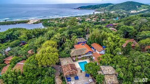 An aerial view of a resort with a swimming pool surrounded by lush greenery, a beach, and a mountainous backdrop. - Pousada Casa do Ceo (Imbituba)