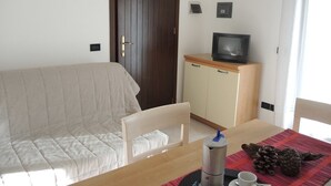 Una habitación con un sofá, una mesa de madera con un mantel individual rojo, un televisor sobre un mueble y una puerta.
