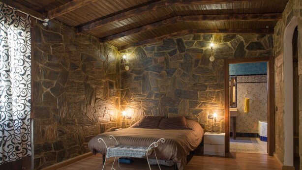 Un dormitorio con pared de piedra, techo de madera y cama con estructura metálica blanca. Hay dos lámparas de noche y una mesita con lámpara.