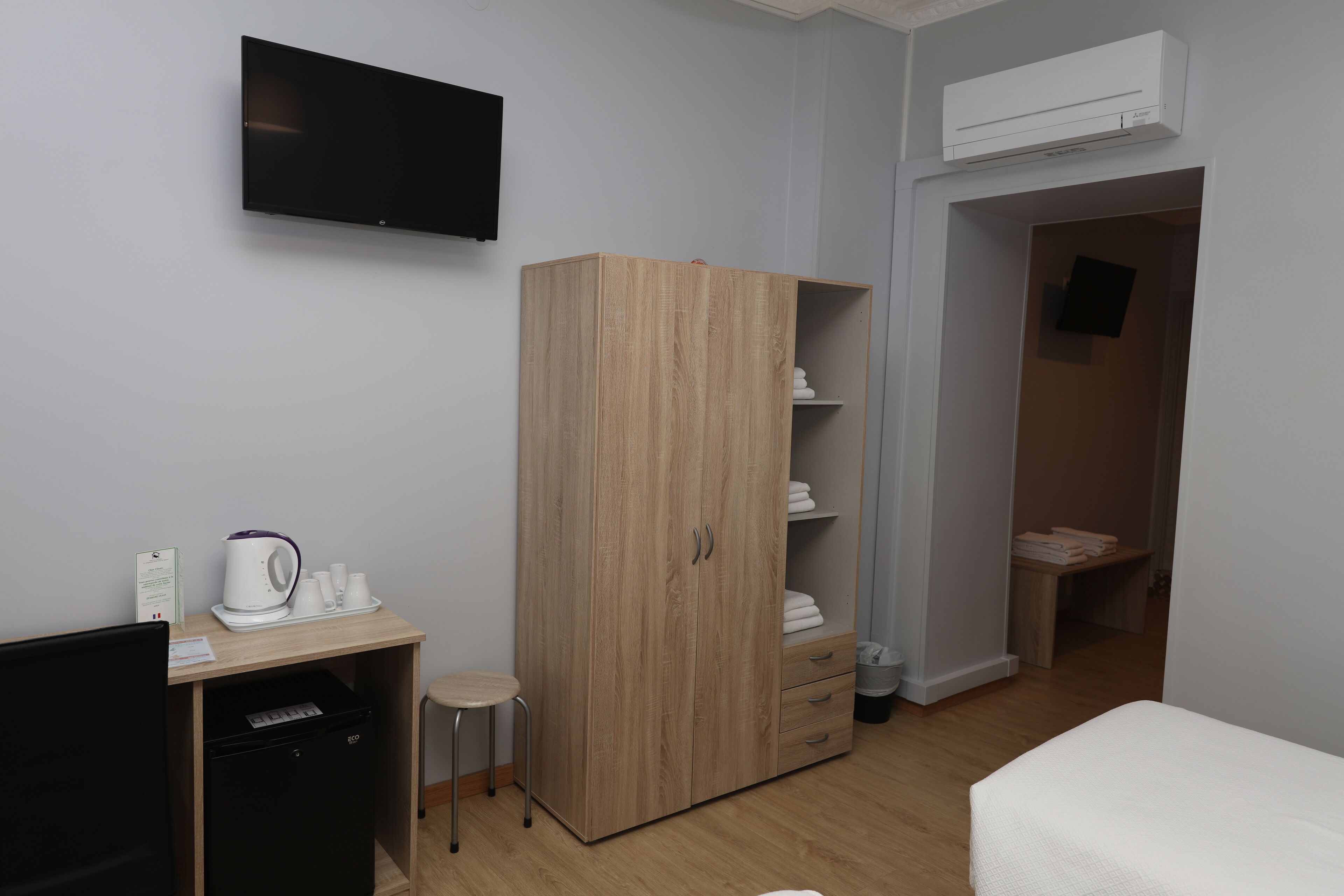 A room with a wooden wardrobe, a desk with a kettle, cups, and a small stool.